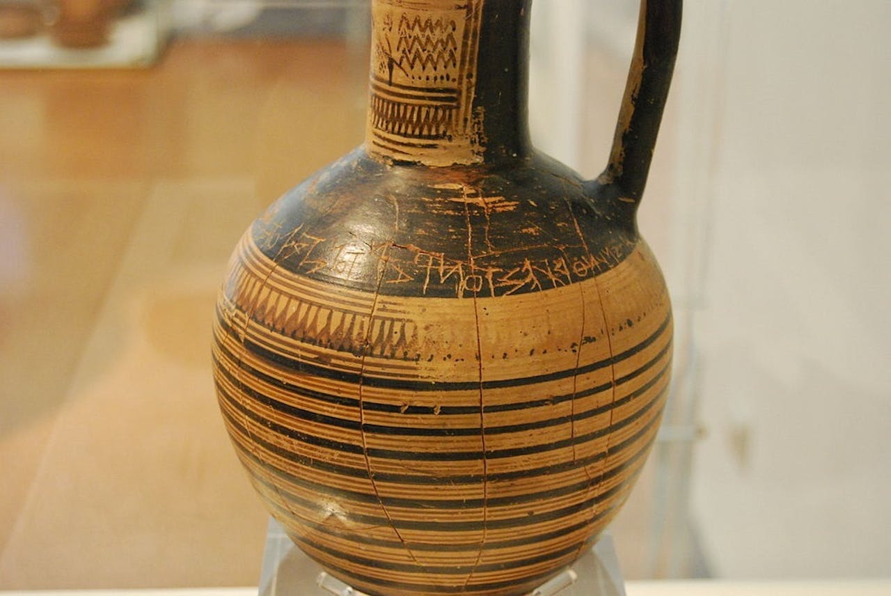 Een van de oudst bekende voorbeelden van het Griekse alfabet is te zien op deze grafvaas, de Atheense Dipylonvaas (circa 740 v. Chr.) Een zwart-bruine vaas tentoongesteld in een museum.