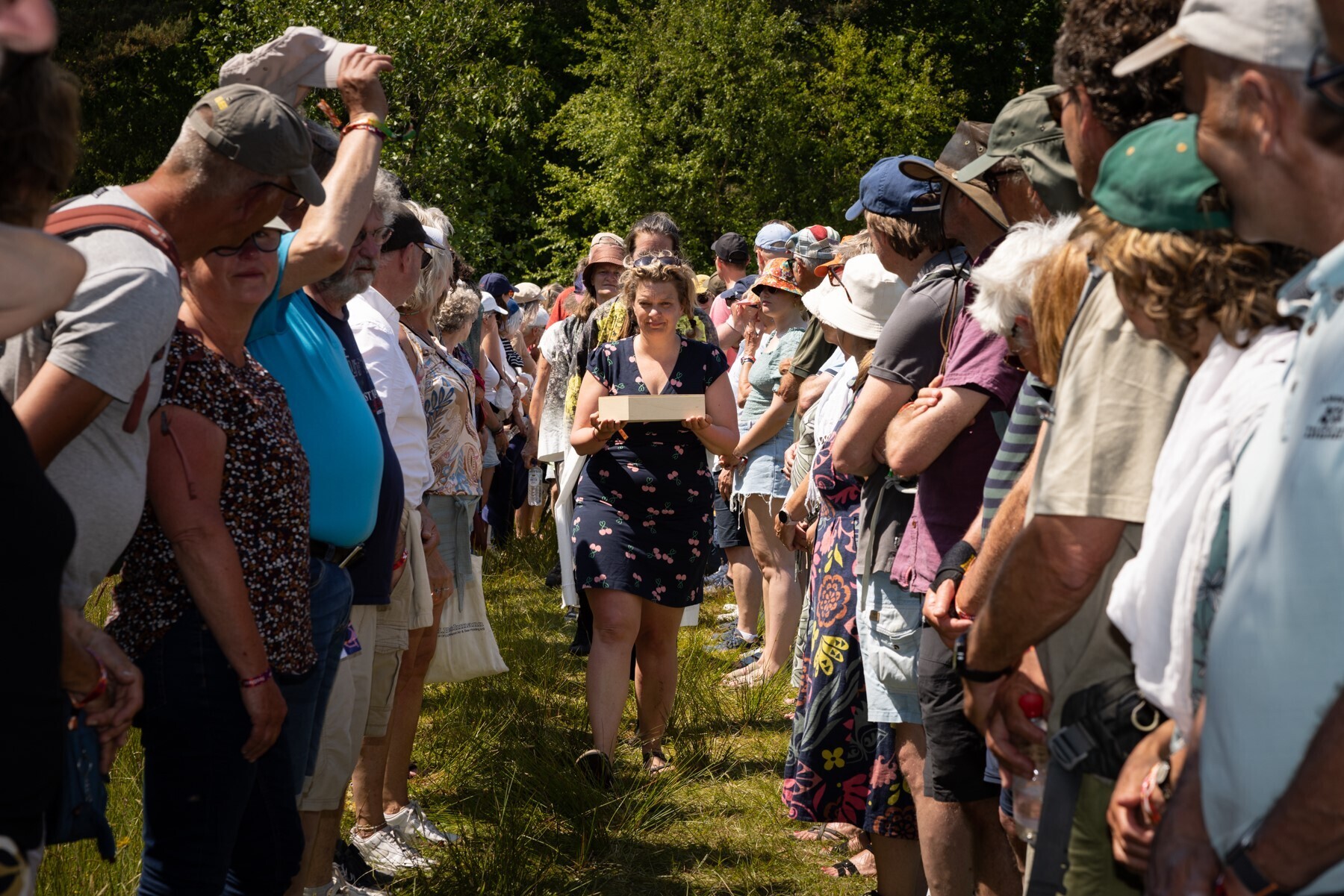 Een erehaag van mensen staat in een grasveld. In de verte loopt een jongedame in een zomerse jurk richting de camera. Ze draagt een houten doos in haar handen.