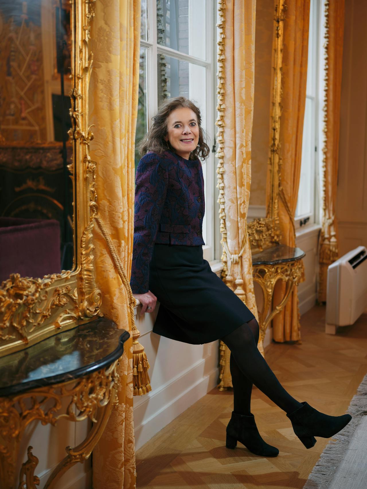 Schrijfster Louise Fresco, een oudere blanke vrouw met donker haar, lacht naar de camera. Ze leunt tegen het raamkozijn van een rijkelijk versierde kamer met gouden lijsten en ornamenten.