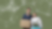 Een jongeman met donker haar en een jongedame met blond haar lachen naar de camera. De jongeman houdt een kartonnen doos vast, de jongedame een plant in een plantenbak.