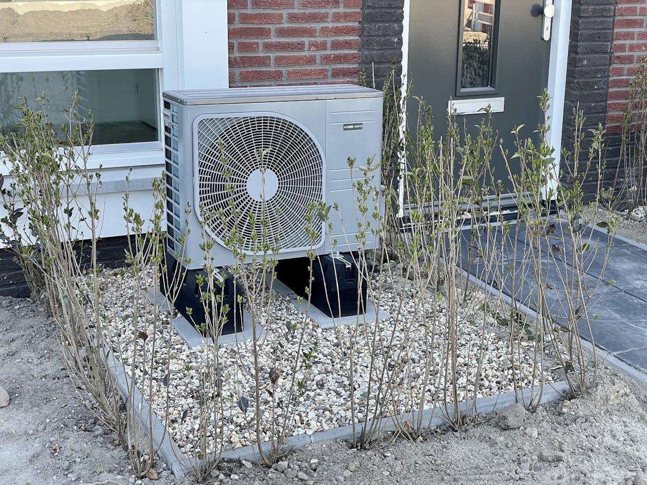 In de voortuin van een huis staat een airconditioning unit.